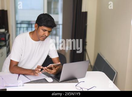 Concentrated young Indian man wearing headphones using laptop computer while working remotely at his home office. Stock Photo