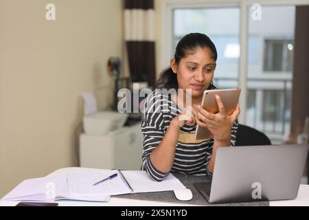 A determined young Indian entrepreneur wearing stylish business casual attire works intently on a digital tablet at her modern home office desk. Stock Photo