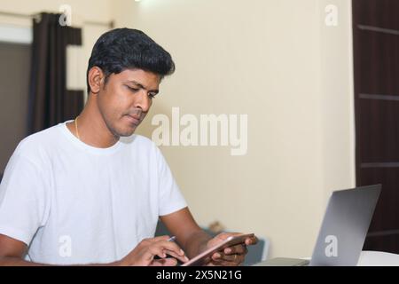 A concentrated young Indian man sits at a desk in his home office, diligently working on a project using both a tablet and a laptop computer. Stock Photo