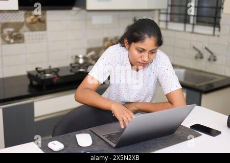 A young Indian woman in casual clothes works diligently on her laptop at a modern kitchen counter Stock Photo