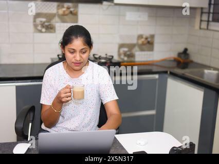 young beautiful and happy Indian woman working at home kitchen with a laptop computer drinking coffee smiling and cheerful in a freelancer job concept Stock Photo