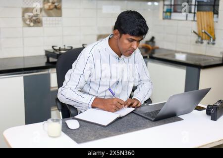 An Indian man concentrates on his work at a desk in his home office. He is using a laptop computer and taking notes in a notebook. Stock Photo