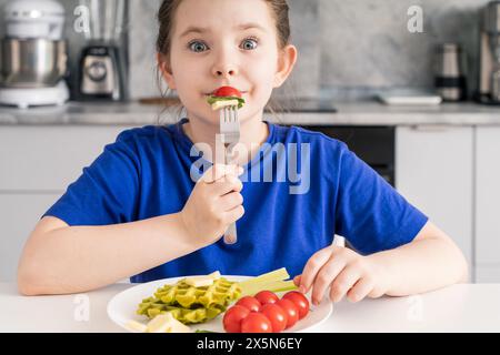 Cheerful little pre-teen girl eating tomato with cheese and herbs in kitchen, waffles and celery on her plate. Proper nutrition for children. selected Stock Photo