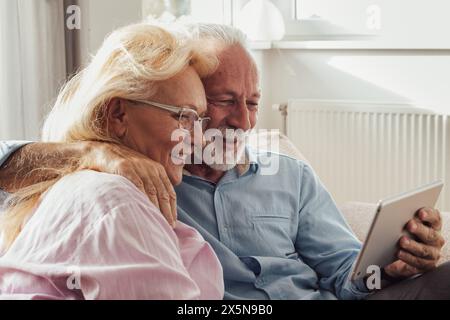 Senior couple hugging on sofa in living room and using tablet for video call with family Stock Photo