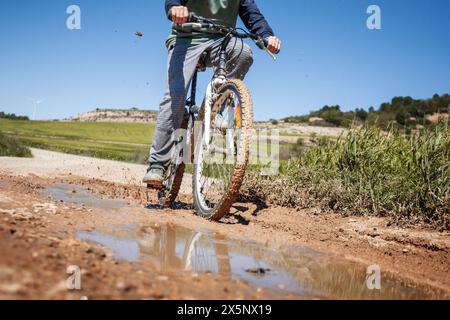 A boy rides his bicycle on a muddy road splashing mud as he passes through a puddle. Stock Photo