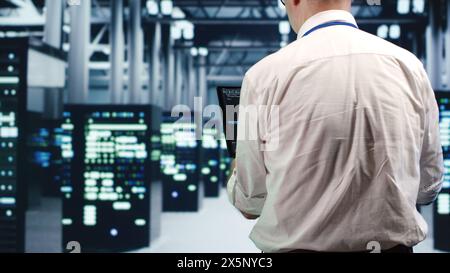 Aged IT consultant in data center, solving tasks on tablet while running scripts on laptop terminal. Trained repairman walking amidst servers, accessing databases and manipulating lines of code Stock Photo