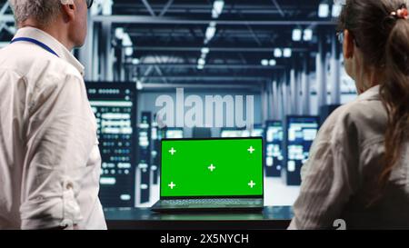 System administrators looking around high tech data center, using mockup laptop to crosscheck disaster recovery plan and assess server hardware in need of replacement, preventing disruption Stock Photo