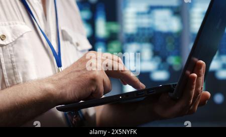Competent IT professional navigating network of supercomputers in industrial server hub. Expert with laptop ensuring optimal operations, doing optimizations and maintenance in data center, close up Stock Photo