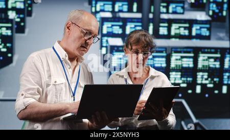 Knowledgeable IT practitioners looking around server farm, using laptop and tablet to crosscheck disaster recovery plan and assess rackmounts in need of replacement, preventing perils Stock Photo