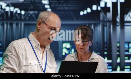 Efficient technical support executives looking around server hub, using laptop to crosscheck disaster recovery plan and assess mainframes in need of replacement, preventing hazards Stock Photo