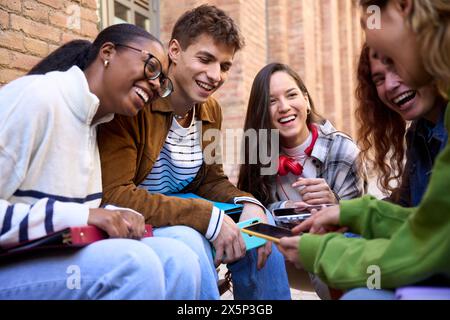 Group of young people students sitting in circle having fun using cell phones together outdoor. Stock Photo