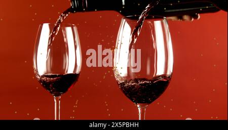 Red wine pouring into two glasses against red background, sparkling droplets visible Stock Photo