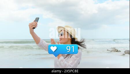 Image of hearts and social media reactions over happy caucasian woman taking photo on beach Stock Photo