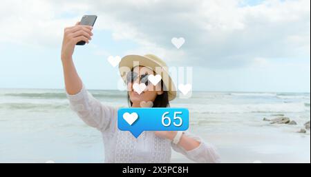 Image of hearts and social media reactions over happy caucasian woman taking photo on beach Stock Photo
