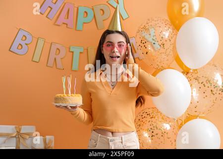 Young woman in party hat with cake celebrating Birthday near orange wall Stock Photo