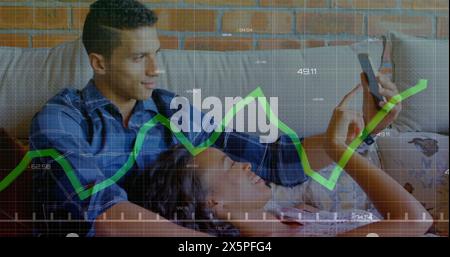 Image of graph and changing numbers, diverse couple watching images on cellphone at home Stock Photo