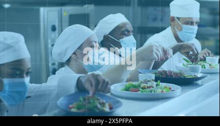 Diverse chefs, with an African American manager, prepare dishes in a pro kitchen. They are wearing white chef uniforms and masks, focusing on plating Stock Photo