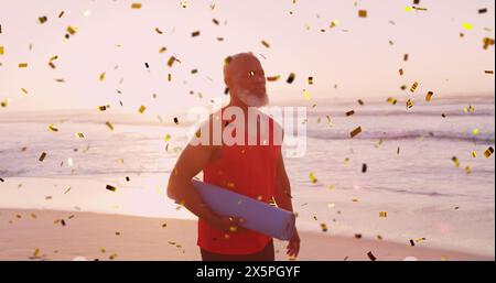 Image of confetti over african american senior man with yoga mat on beach at sunset Stock Photo