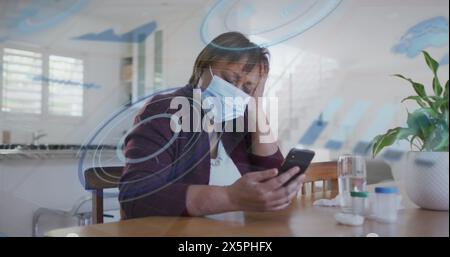 Biracial woman wearing mask, sitting at table, looking at smartphone Stock Photo