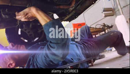 A mechanic wearing blue overalls is fixing car from underneath Stock Photo