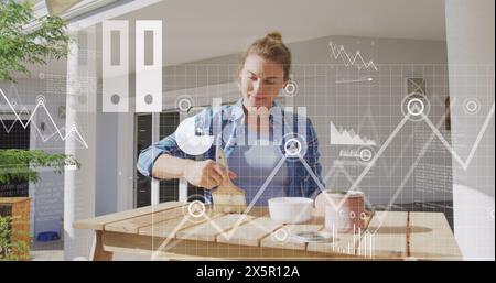 Image of multiple icons, graphs, map, caucasian woman painting wooden table at home Stock Photo