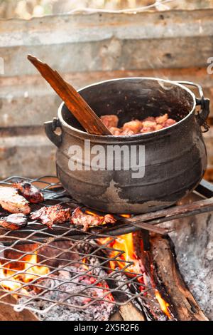 african cooking , meat on the barbeque, large black cauldron pot boiling with stew in the background Stock Photo