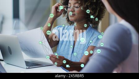 Image of globe of icons over two diverse female office colleagues discussing over a document Stock Photo