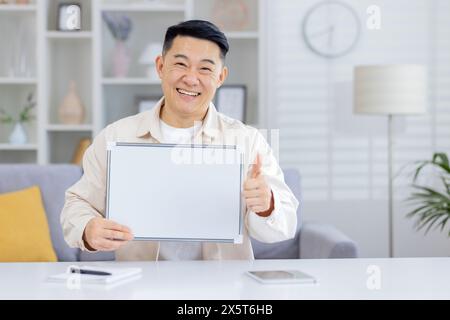 Asian man confidently presents a blank whiteboard while giving a thumbs-up. Positioned in a well-lit home office, this image perfectly captures a moment of positive communication, Stock Photo
