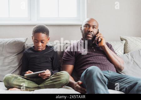 Father using phone sitting on sofas next to son (10-11) using digital tablet Stock Photo