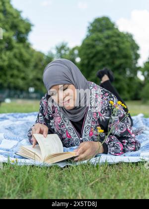 UK,Sutton,Woman in headscarf reading book on lawn in park Stock Photo