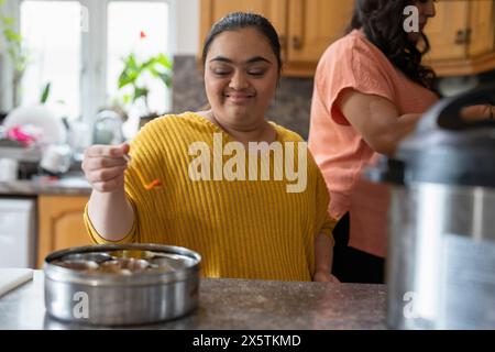 Woman with down syndrome adding spices while cooking with mother in kitchen Stock Photo