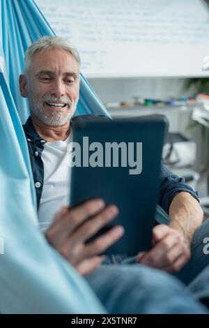 Smiling mature man lying in hammock and using digital tablet Stock Photo