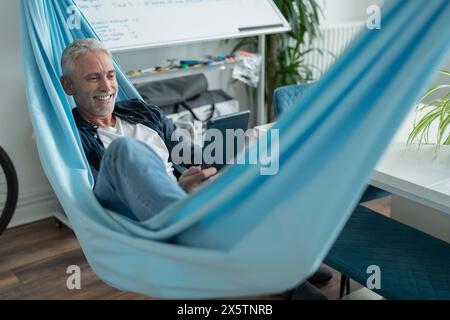 Smiling mature man lying in hammock and using digital tablet Stock Photo