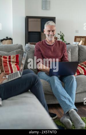 Smiling male couple working on laptops on sofa at home Stock Photo