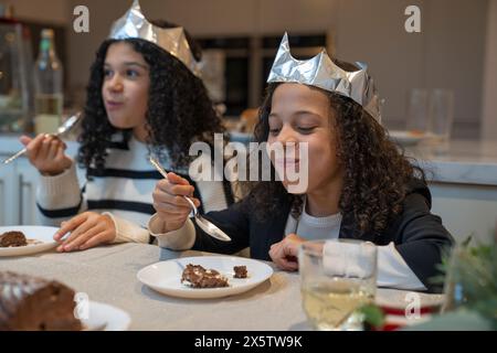 Sisters in paper crowns eating chocolate Yule log cake at Christmas dinner table Stock Photo