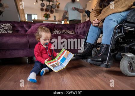 Baby boy ¾playing with book next to mother on wheelchair Stock Photo