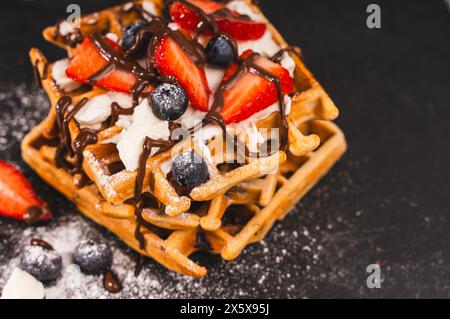 Dessert close-up. Waffles with cottage cheese cream, strawberries and blueberries, sprinkled with powdered sugar and drizzled with chocolate on a blac Stock Photo