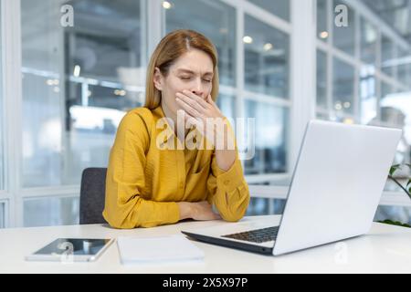 A tired young adult woman in a yellow shirt, yawning as she works on her laptop in a brightly lit office. Stock Photo