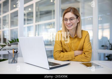 A young professional woman with glasses smiles as she works on her laptop in a bright, modern office environment. Stock Photo