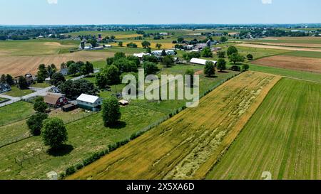 Aerial photograph showcases a mixed landscape of residential and agricultural areas, featuring homes, farm buildings, and variously colored fields under a clear blue sky. Stock Photo