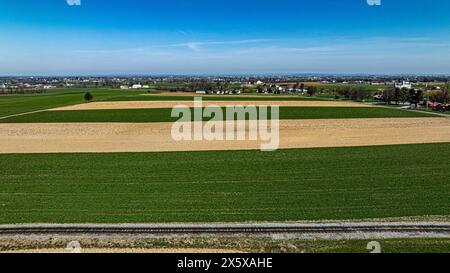 A stunning aerial shot capturing the vibrant patchwork of farmland fields alongside a small village, with distinct patches of green and brown fields under a clear blue sky. Stock Photo