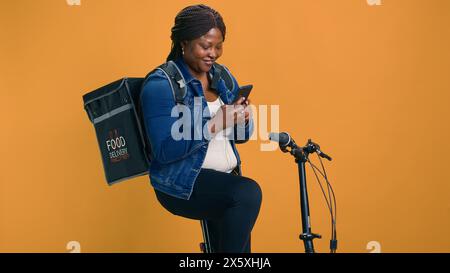 Healthy african american female with bag on her back using mobile communication for on-the-go urban delivery service. Active black woman happily receiving online orders on cell-phone from customers. Stock Photo