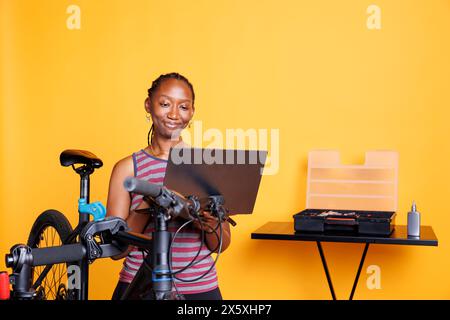 In front of yellow background, black woman performs bike maintenance by utilizing digital laptop for instructions. African american female using minicomputer and professional tools for fixing bicycle. Stock Photo