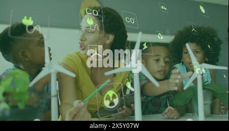 Image of energy icons over happy diverse female teacher and pupils with wind turbines in class Stock Photo