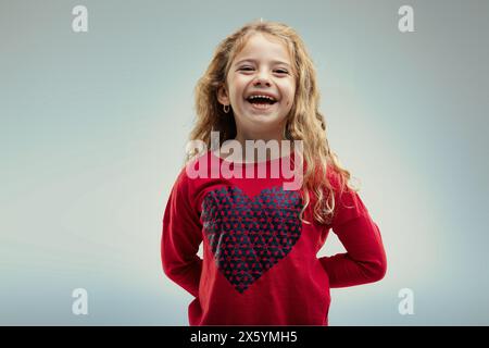 Young girl with curly blonde hair laughs joyously, wearing a red sweater adorned with a large heart pattern, her happiness contagious Stock Photo