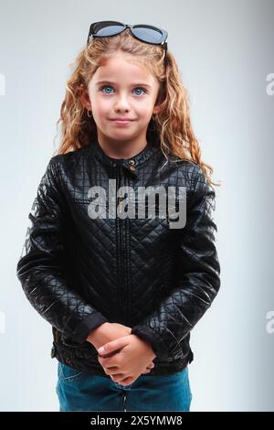 Young girl with blue eyes and curly blonde hair, wearing sunglasses on her head and a stylish black leather jacket, clasps hands together Stock Photo