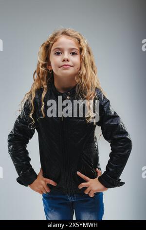 Young girl with curly blonde hair stands confidently, hands on hips, wearing a quilted black jacket and blue jeans against a grey background Stock Photo