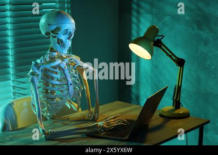 Waiting concept. Human skeleton sitting at wooden table with laptop indoors Stock Photo