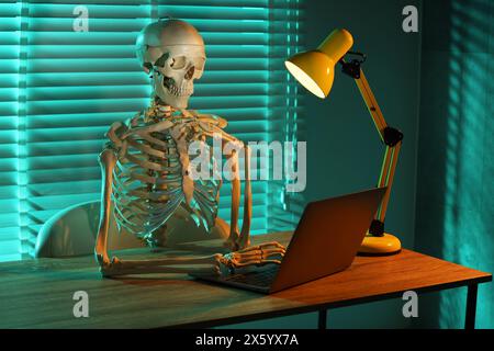 Waiting concept. Human skeleton sitting at wooden table with laptop indoors Stock Photo