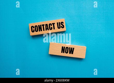 Contact us now words written on wooden blocks with blue background. Conceptual contact us now symbol. Copy space. Stock Photo
